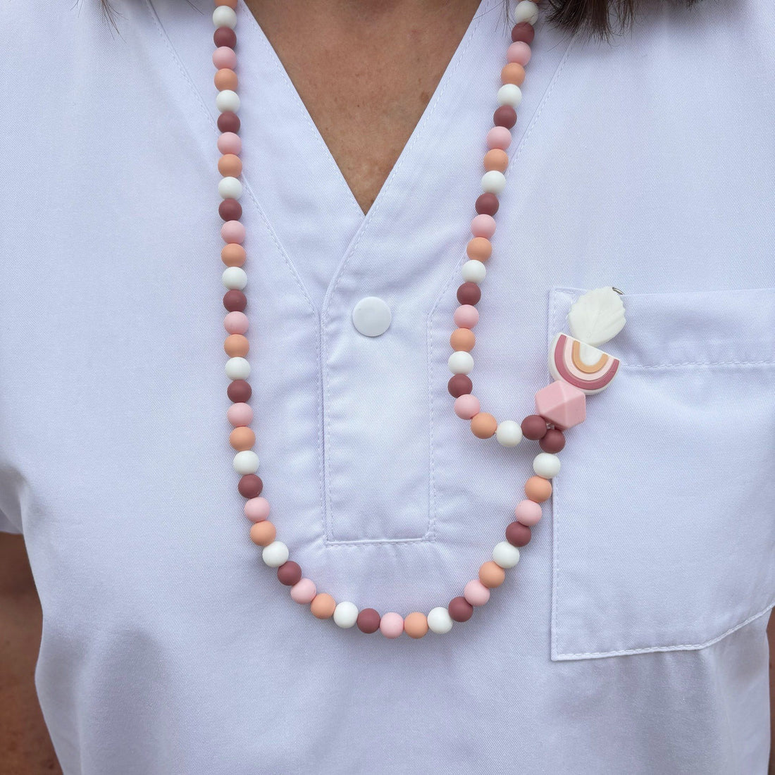 Porte-badge en perles silicone roses, pêches, terracotta et blanches, porté autour du cou sur une blouse blanche, avec en pendentif une feuille blanche, un arc enc ciel dans les tons de roses, pêche et une perle hexagonale rose quartz