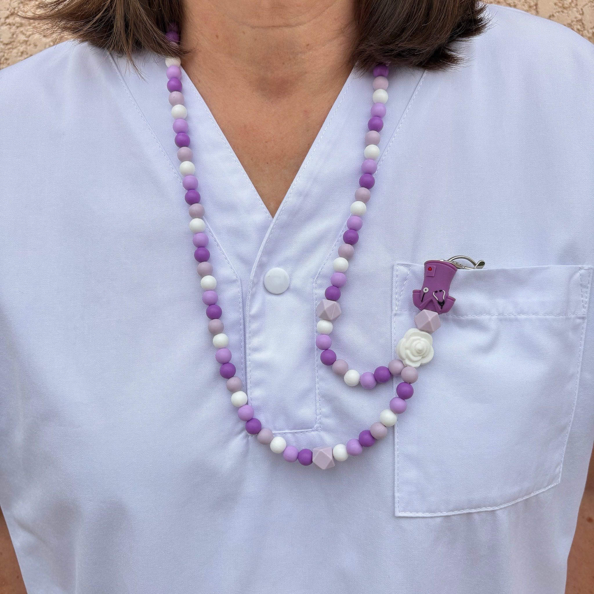 Porte badge tour de cou en perles de silicone blanches, parme, lilass, violet avec en pendentif une fleurs blanche, un cube hexagonale parme et une chemise d'infirmière violette